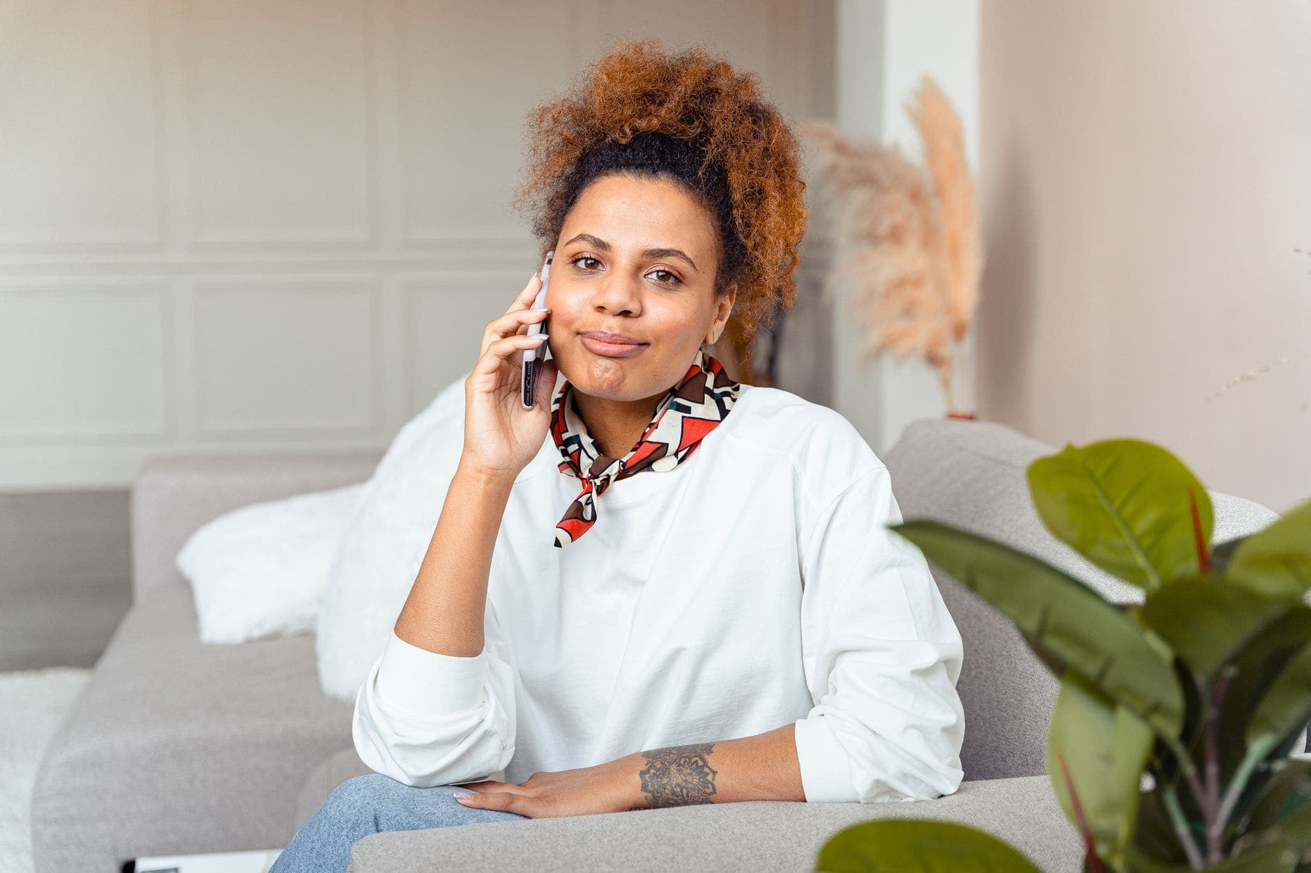 A woman in a white blouse holds a phone to her ear while sitting on a grey sofa.