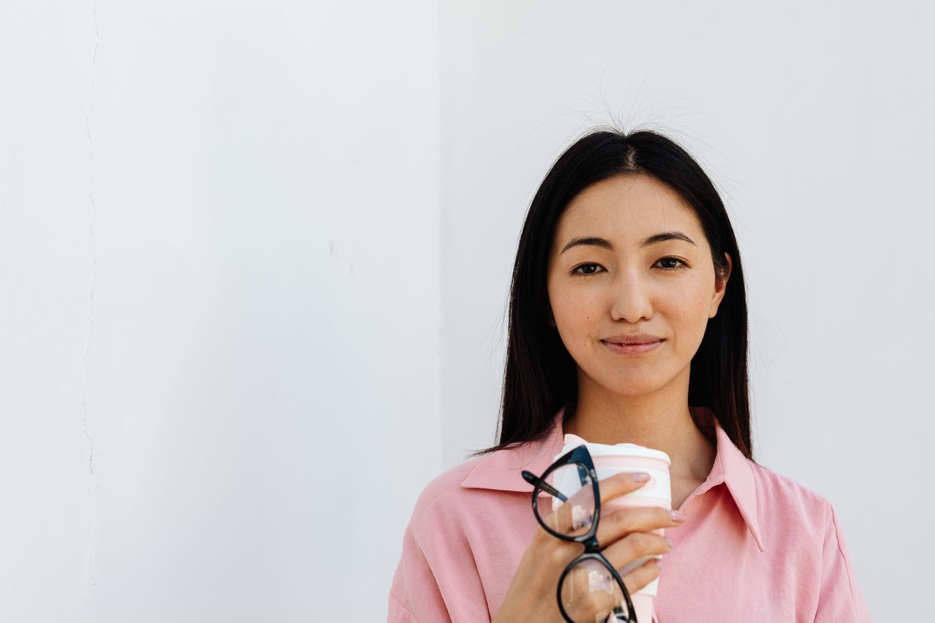 A woman in a pink shirt holds a takeaway cup and glasses against a pale wall.