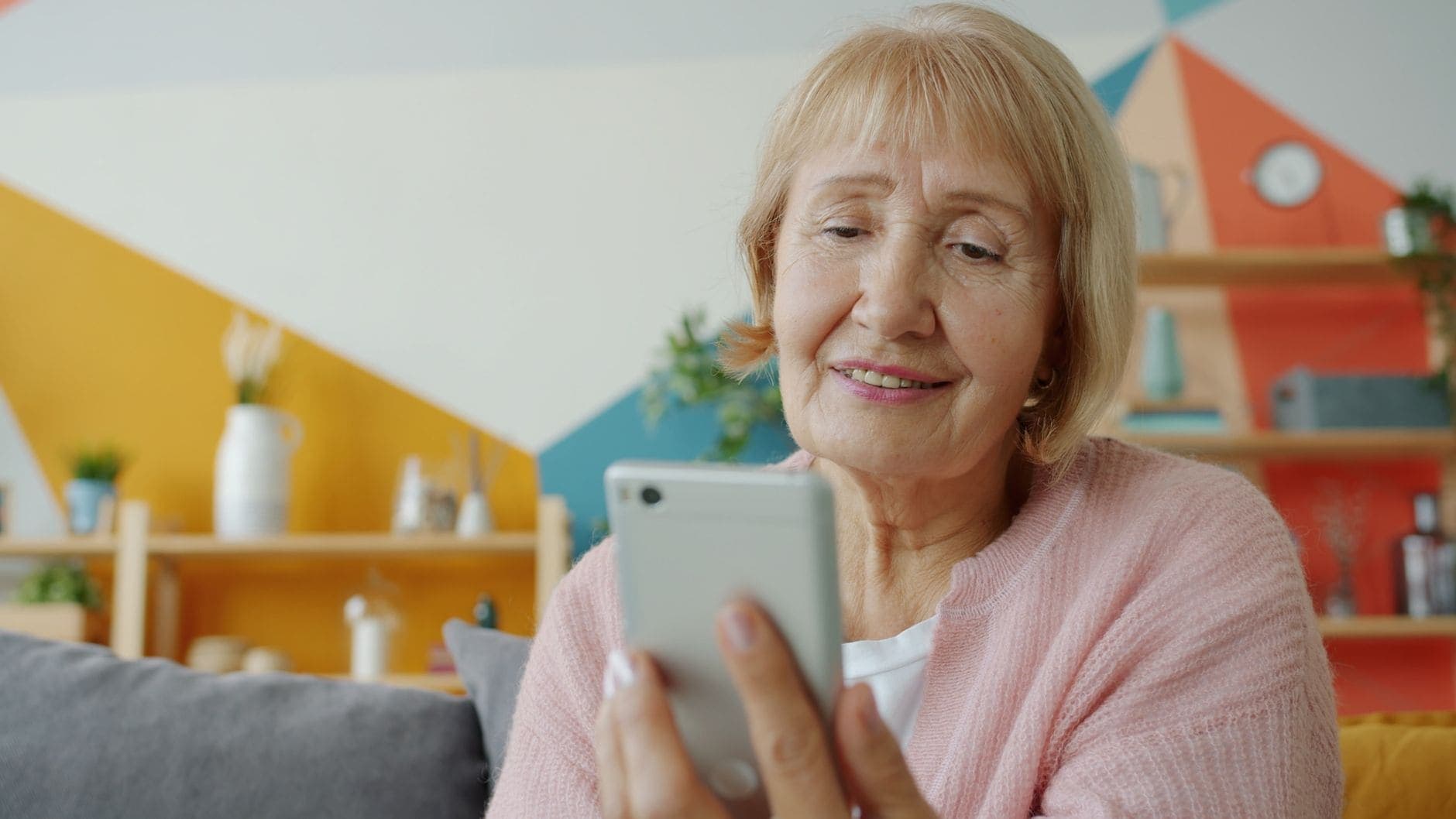 Rózsaszín kardigánt viselő nő mosolyogva nézi a telefonját egy színes fal előtti kanapén.