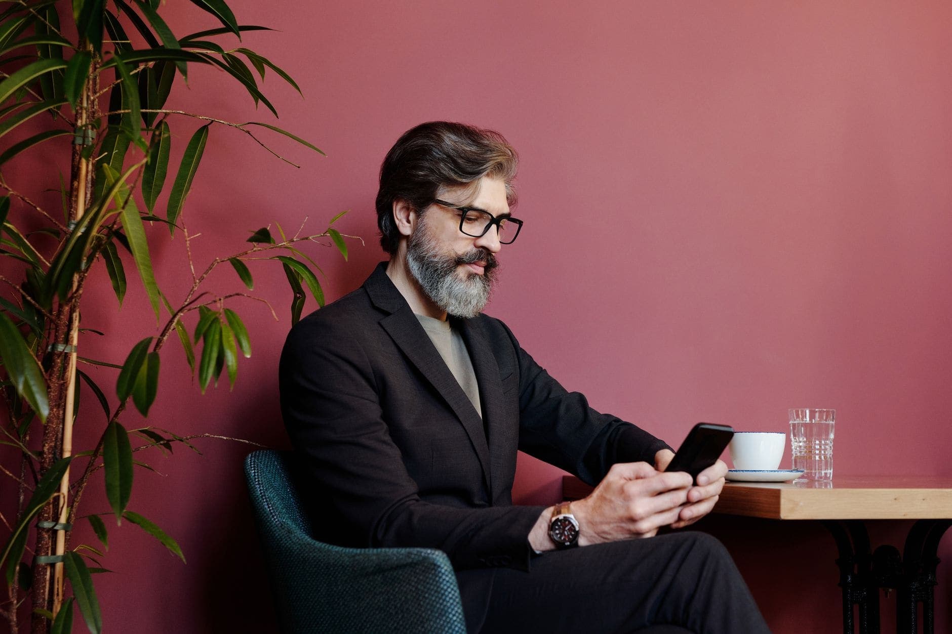 Um homem de barba e óculos olha para o celular à mesa de um café, diante de uma parede vermelha.