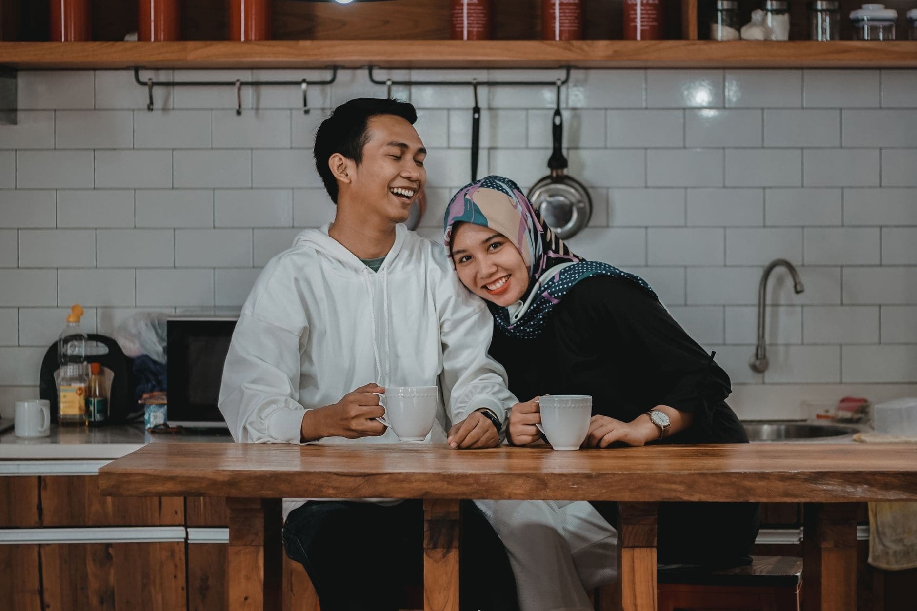 A young couple laugh together over mugs of coffee at a bright tiled kitchen counter