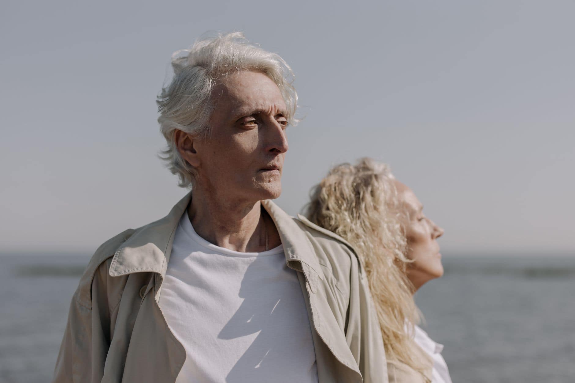 Senior couple standing by the sea on an overcast day, both looking into the distance.