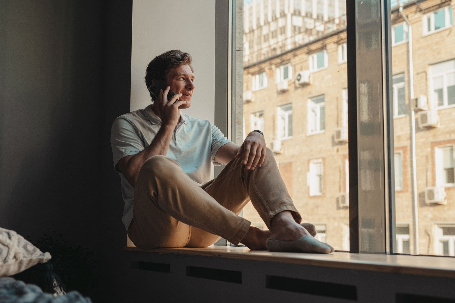 Egy férfi az ablakpárkányon ülve mosolyog, és a füléhez tartja a telefonját.