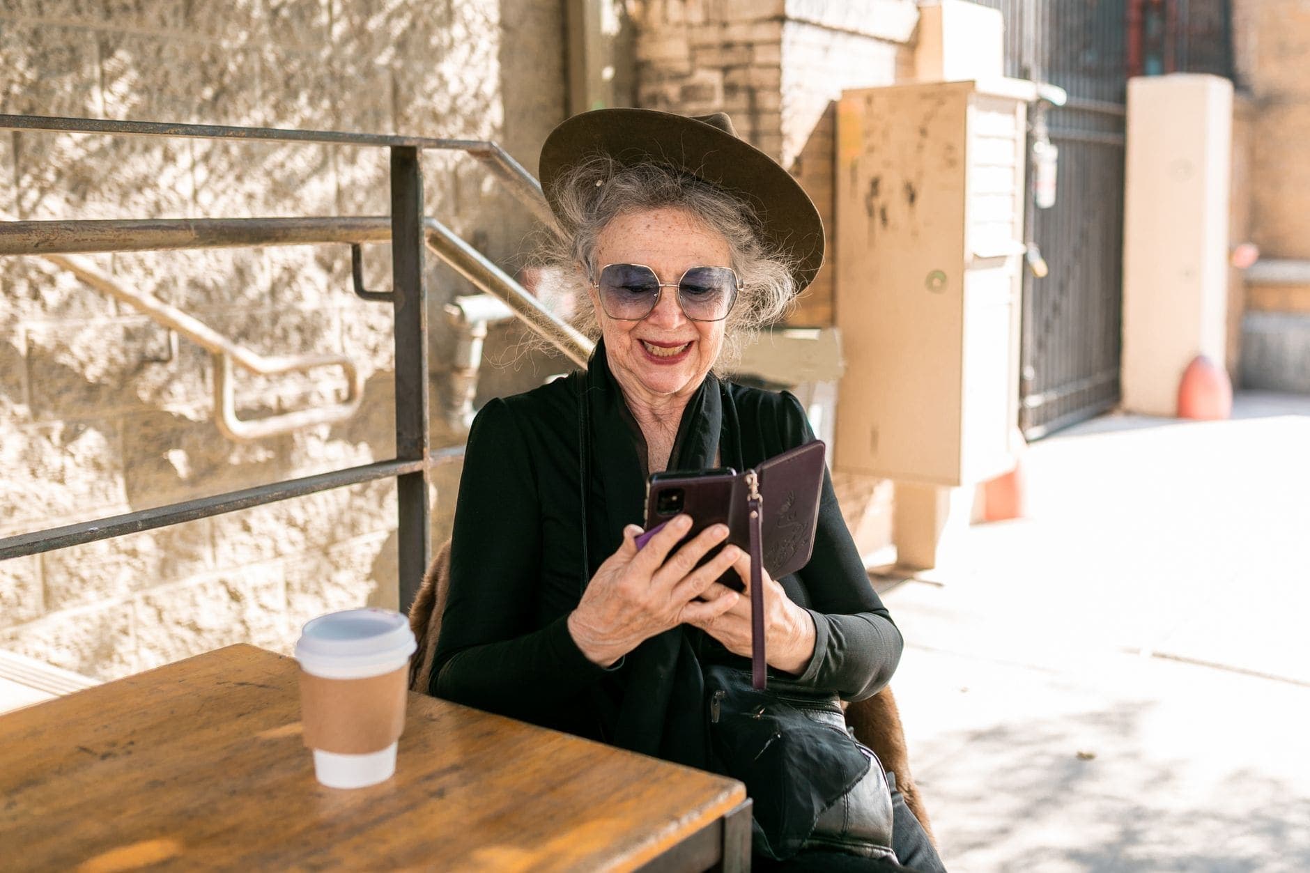 A woman in a black hat smiles at her phone at an outdoor café table beside a takeaway cup.
