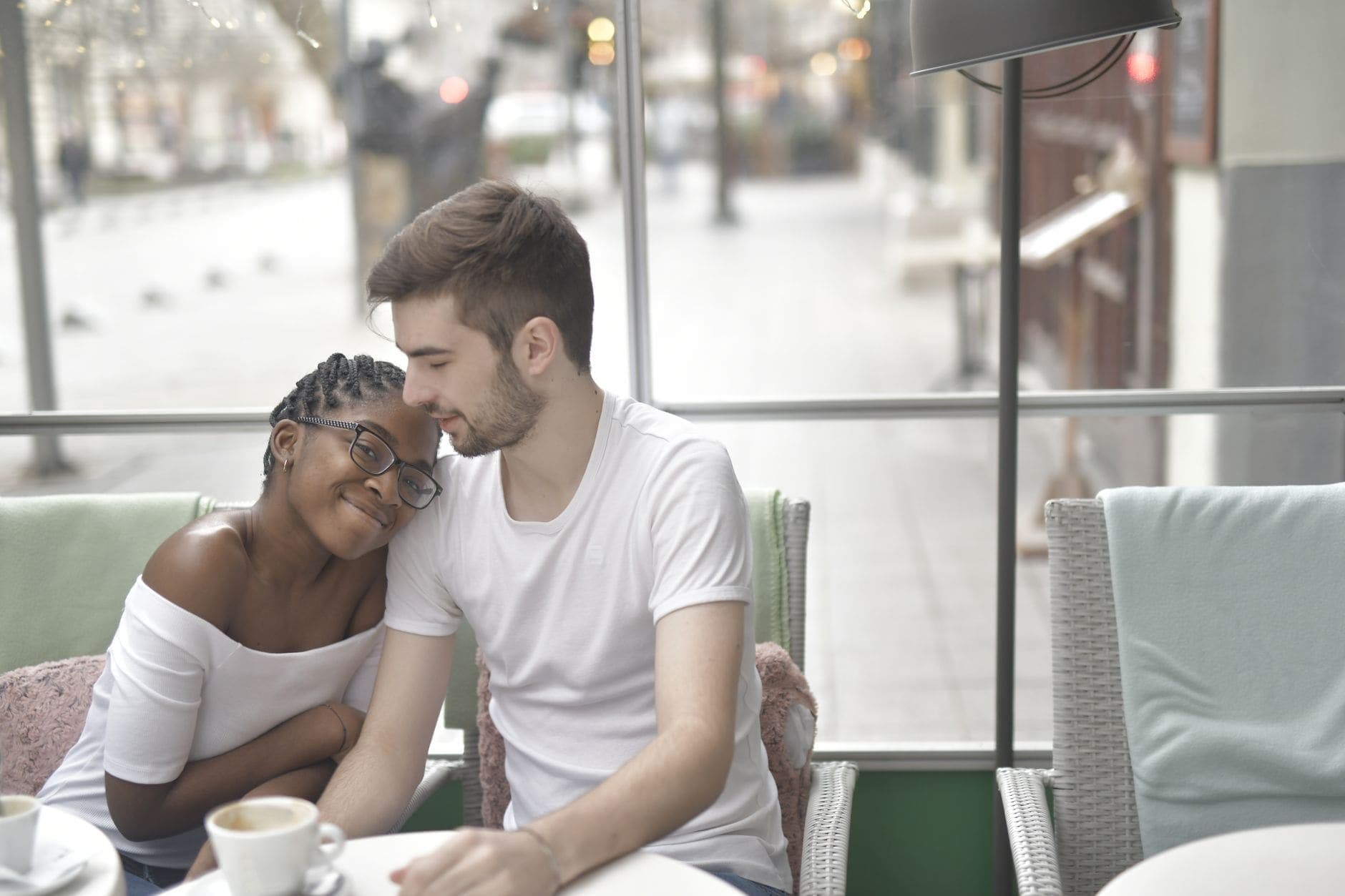 A woman with glasses rests her head against a man beside a bright café window.