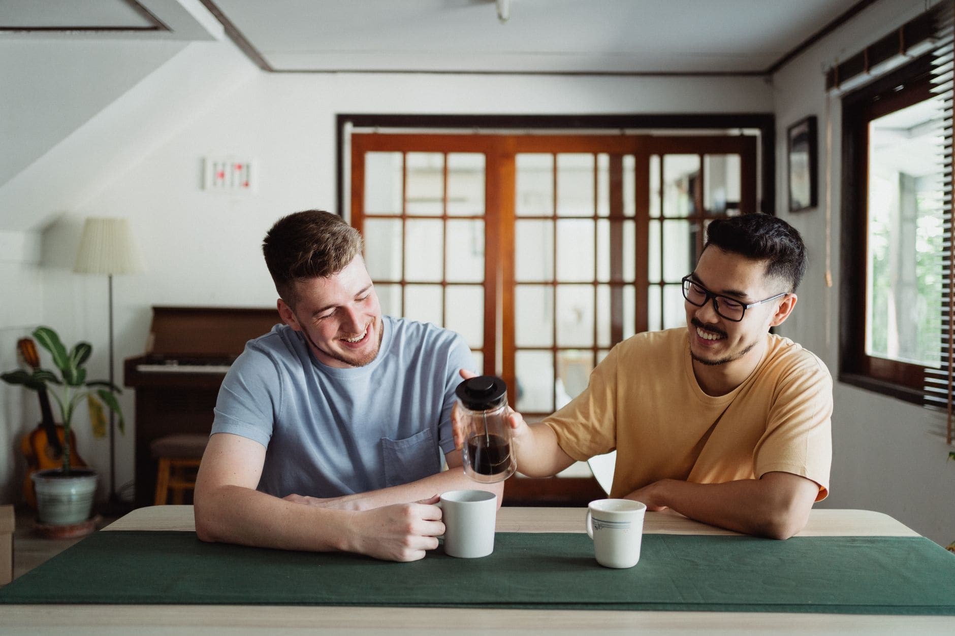 Two men smile at a table as one holds a cafetière above a mug in a bright room.