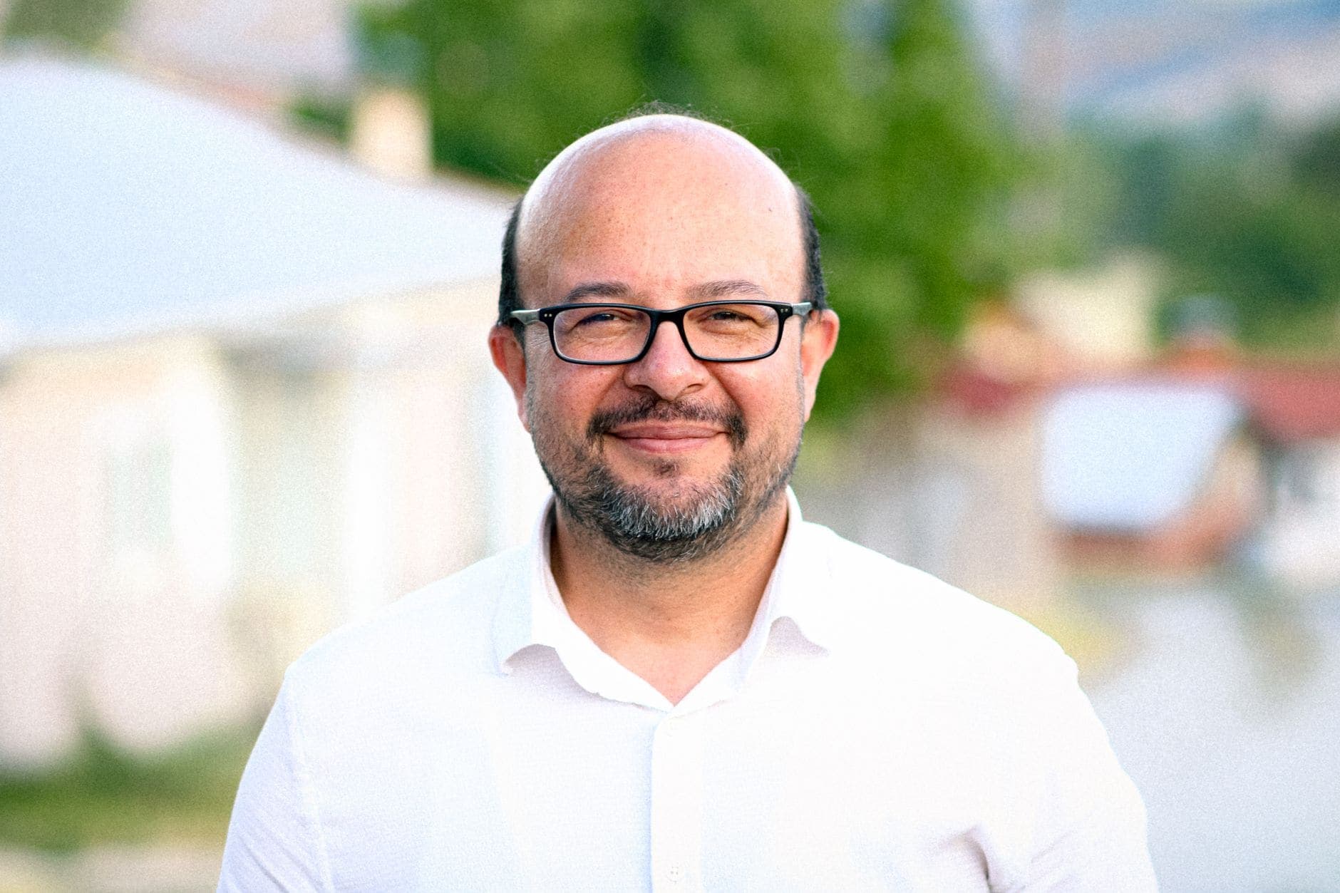 Szemüveges, őszülő szakállú férfi fehér ingben mosolyog az elmosódott zöld háttér előtt.