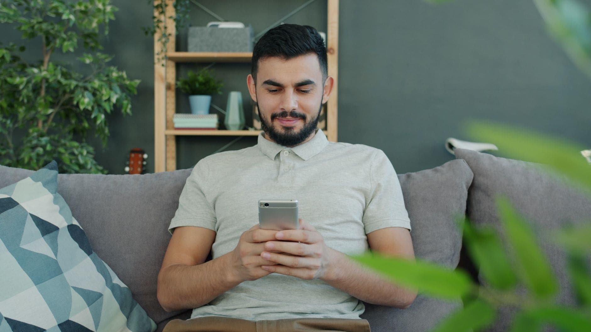 Szakállas férfi nézi a telefonját egy szürke kanapén, mögötte növények és egyszerű polc látható.