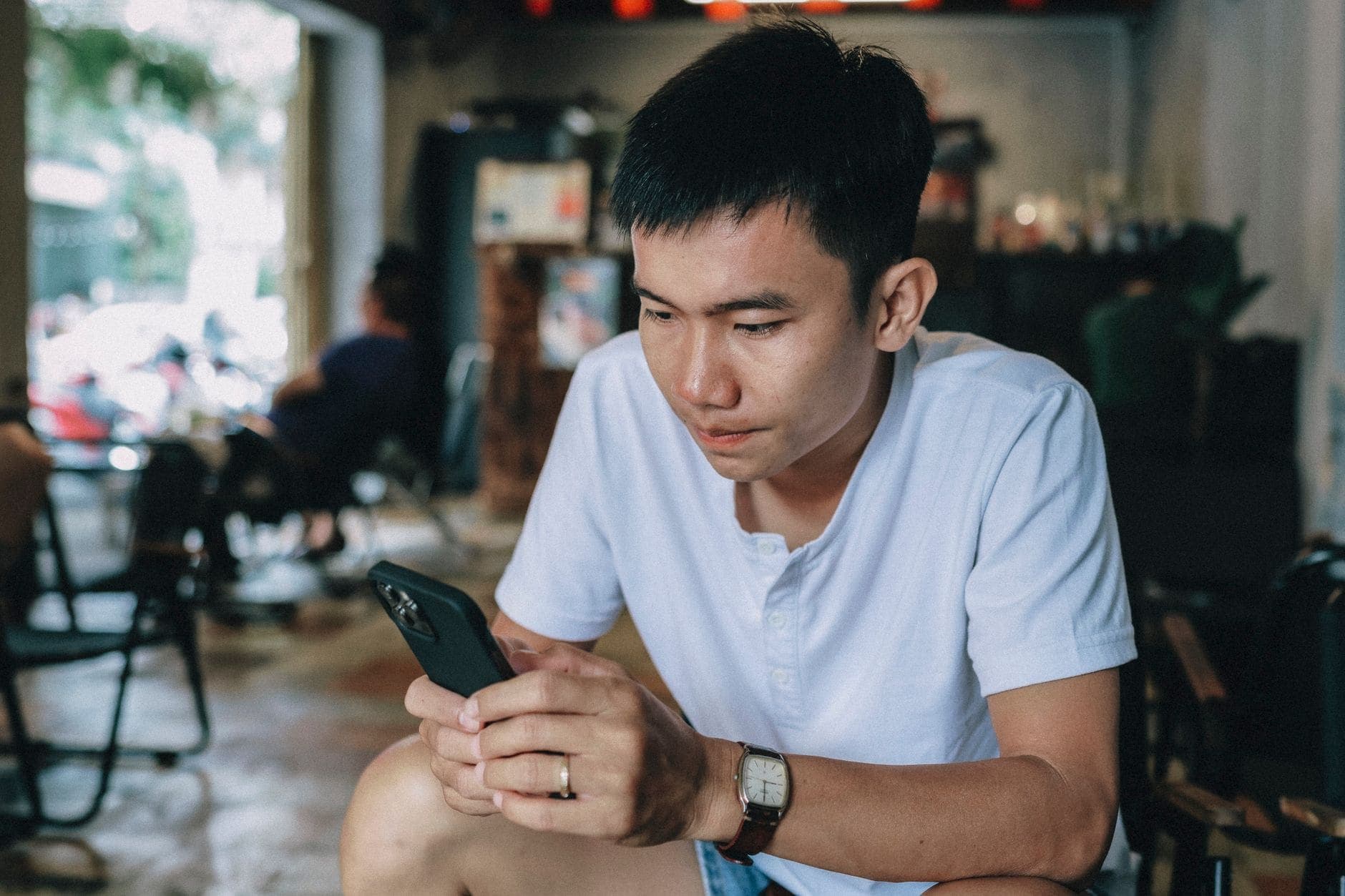 A man in a white polo reads his phone at a café table, with the street soft in the background