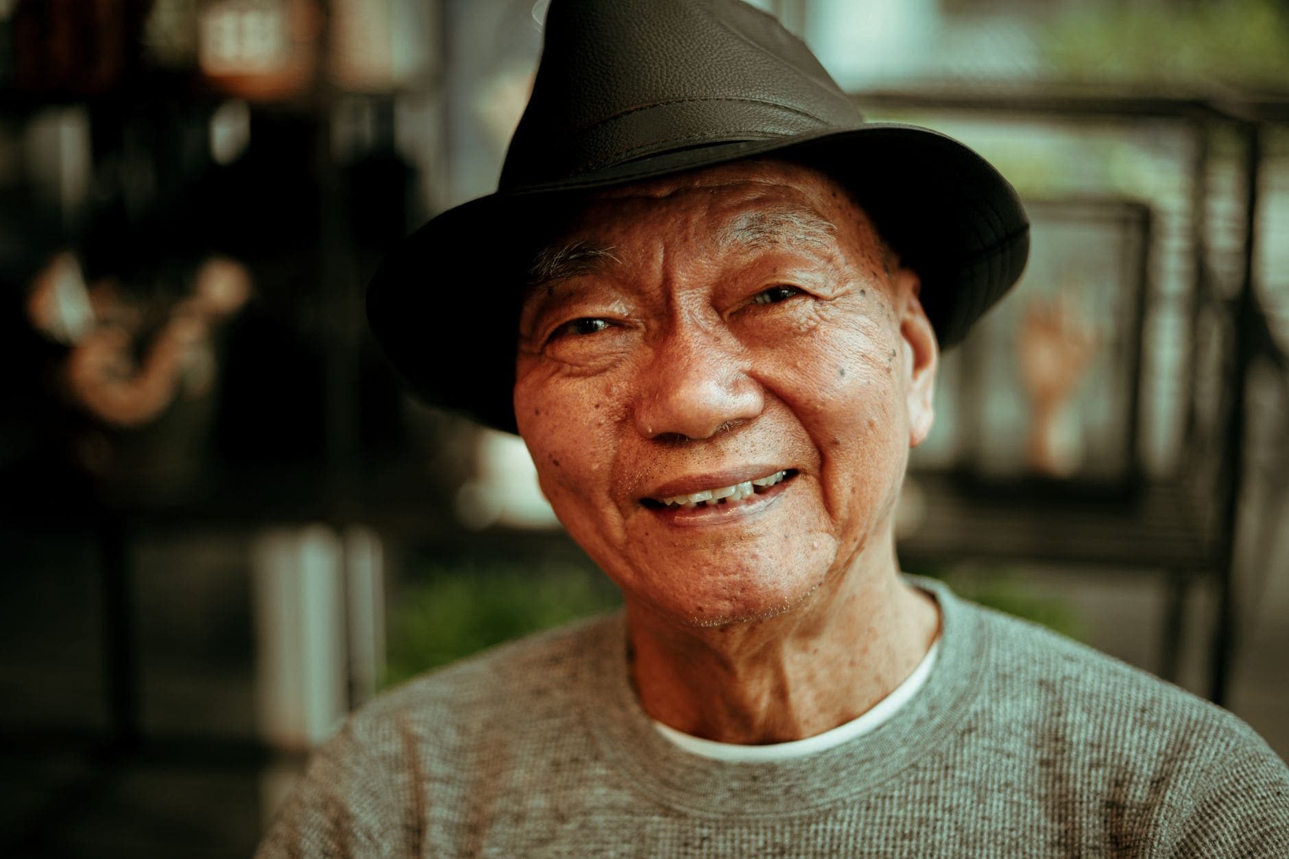 An older man in a dark hat and a grey jumper smiles in a close-up portrait.