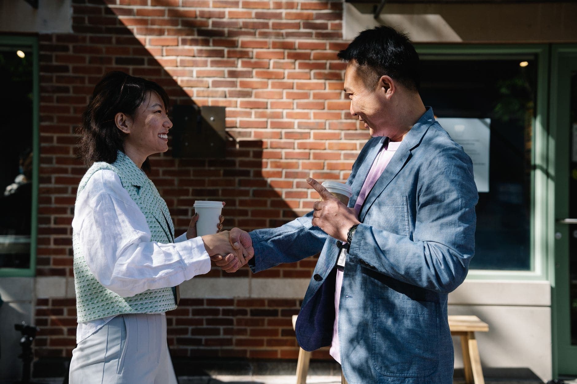 A woman and a man in smart casual clothes shake hands with takeaway coffees outside a sunlit brick cafe