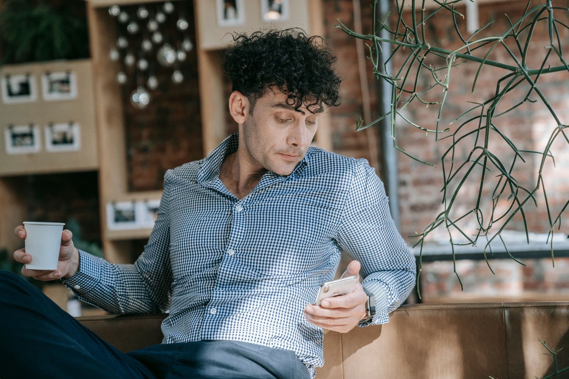 Um homem de camisa aos quadrados olha para o telemóvel enquanto segura um copo.