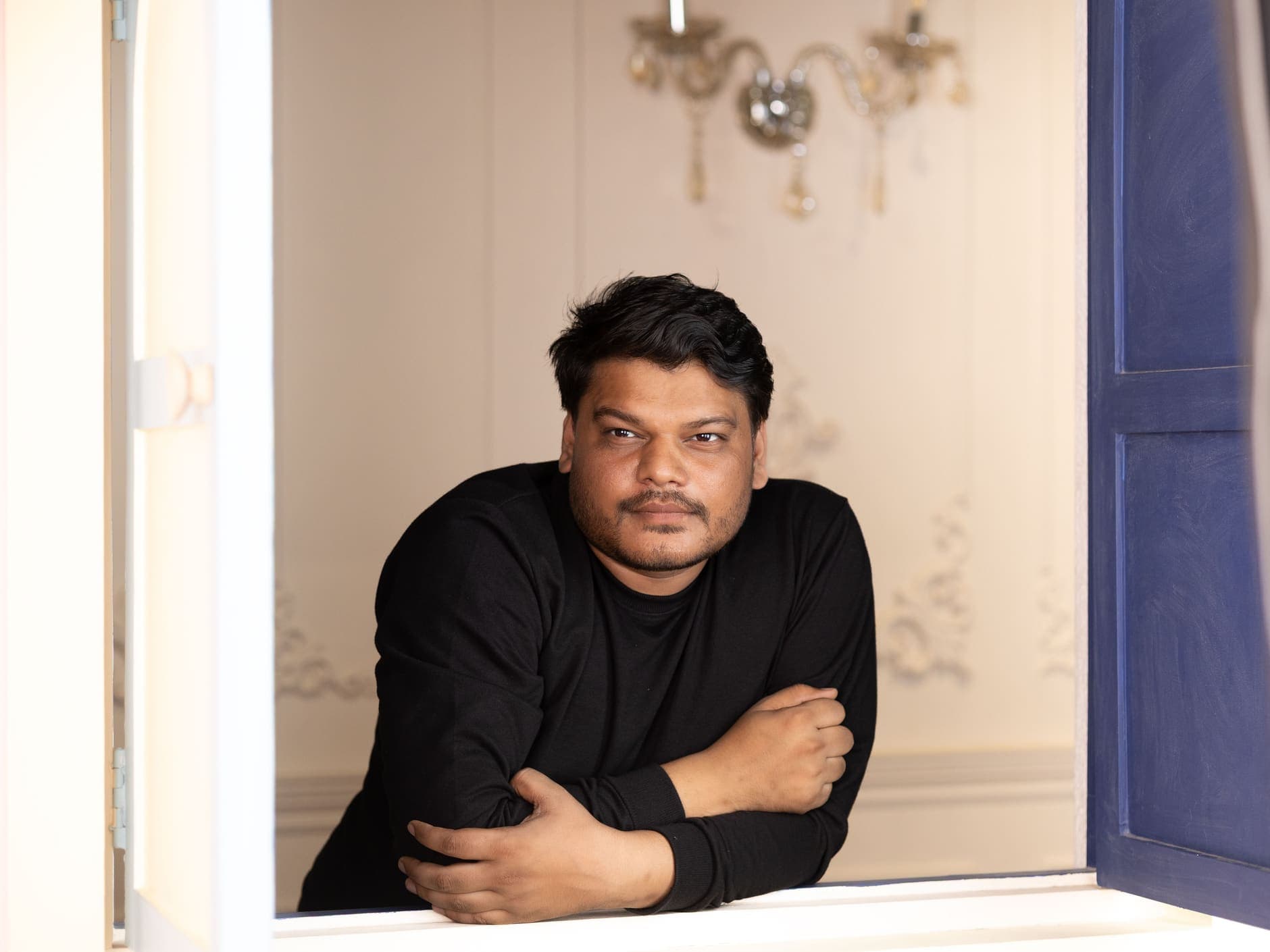 A man in a black top rests his arms on a white windowsill beside a blue shutter.