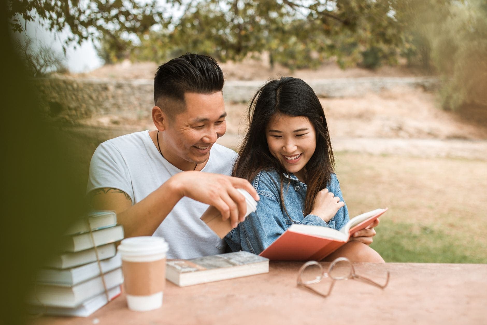 A man and a woman laugh together over an open book and coffee at an outdoor table.