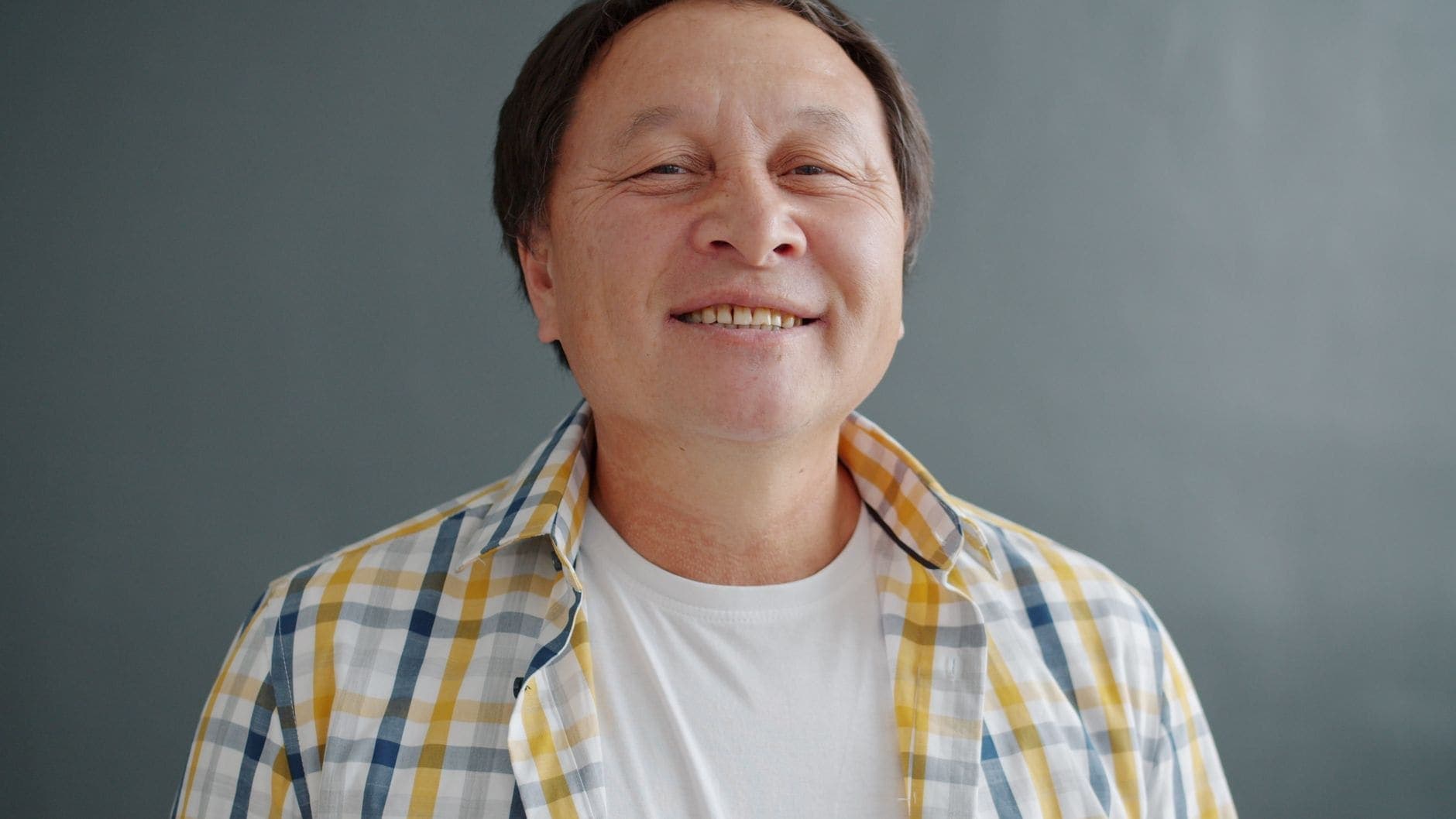 A man in a yellow and grey checked shirt smiles against a plain grey background.
