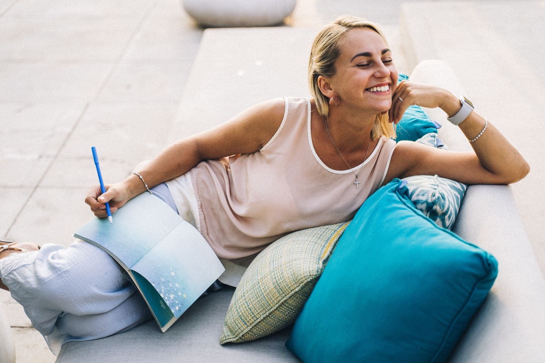 A woman smiles beside turquoise cushions with an open notebook on her lap.