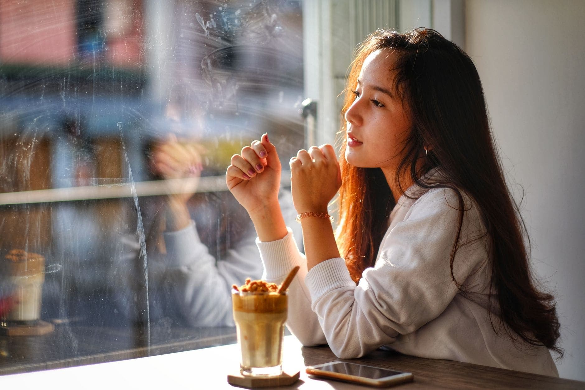 A woman in a white top looks out of a café window, with a drink and phone on the table.