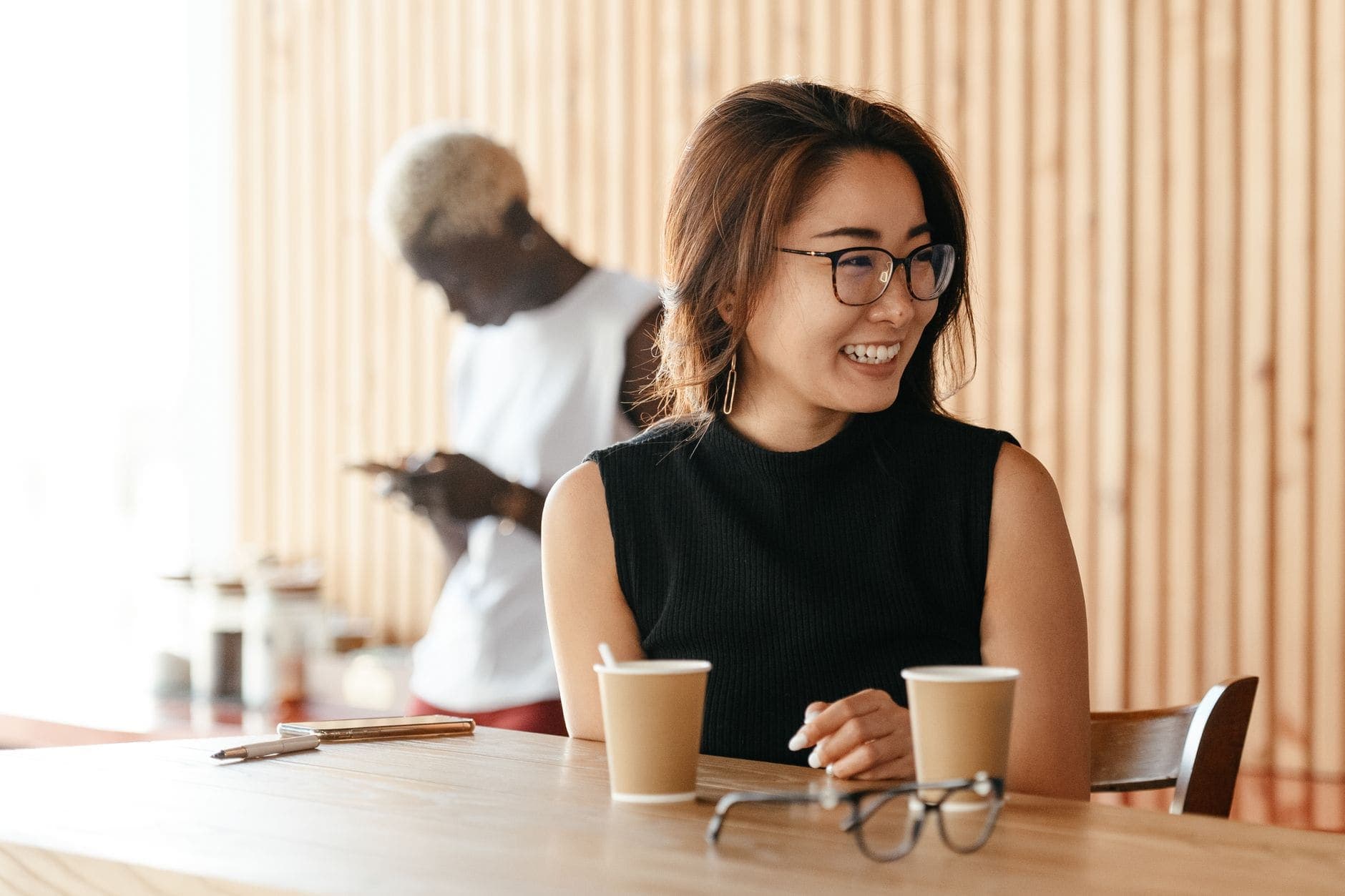 A woman with glasses smiles to one side at a café table with two paper cups.