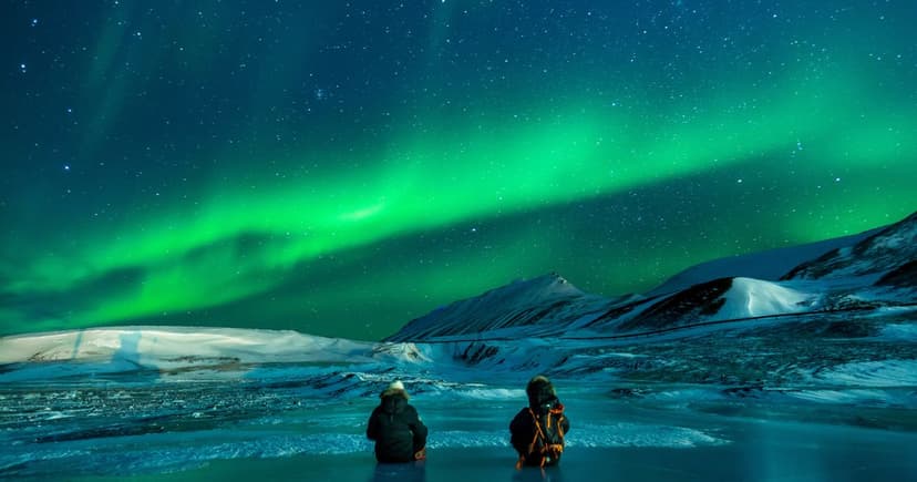 Kaksi ihmistä istuu jäällä ja katselee vihreitä revontulia lumisten tunturien yllä.