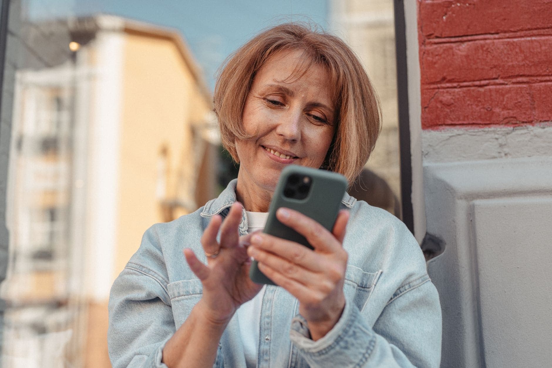 A woman in a pale denim shirt smiles while looking at the phone in her hands.