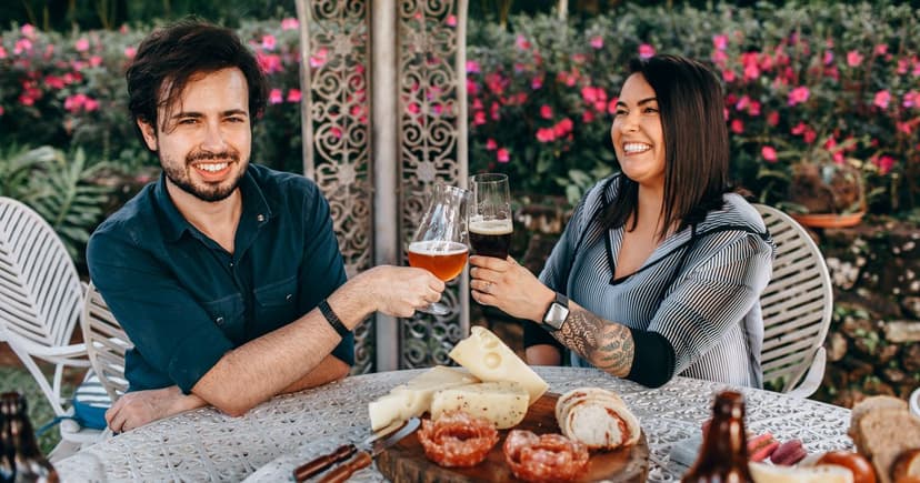 A smiling man and woman touch glasses across a patio table set with cheese and bread, flowering shrubs behind them.