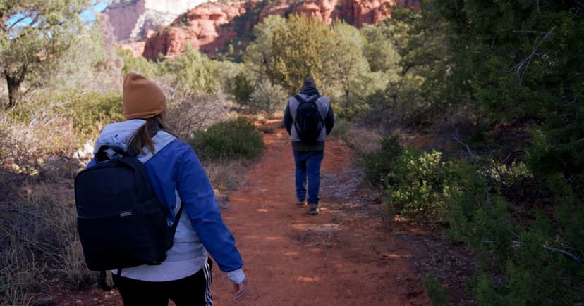 Two hikers in jackets and a knit hat follow a red dirt trail through juniper scrub toward tall sandstone cliffs.