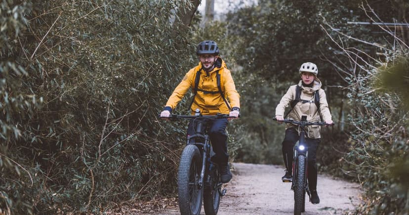 A man in a yellow rain jacket and a woman in a beige jacket ride bikes side by side along a narrow gravel trail through winter woodland.