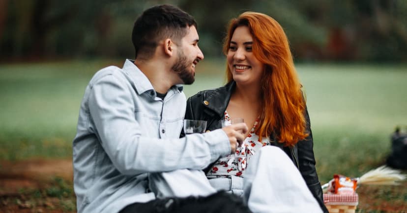 A man in a pale denim jacket and a red-haired woman in a black leather jacket sit close together on the grass in a green park, laughing over small glasses of wine.