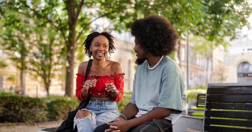 A smiling couple sit talking on a bench in a leafy city park on a sunny afternoon.