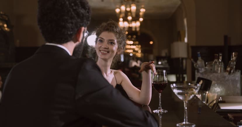 A well-dressed couple talk over a glass of red wine and a cocktail at a chandelier-lit hotel bar.