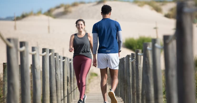 A smiling woman in a grey vest and burgundy leggings walks down a wooden beach path toward the camera as a man in a navy T-shirt and white shorts heads the other way, low post fences and pale dunes on either side.