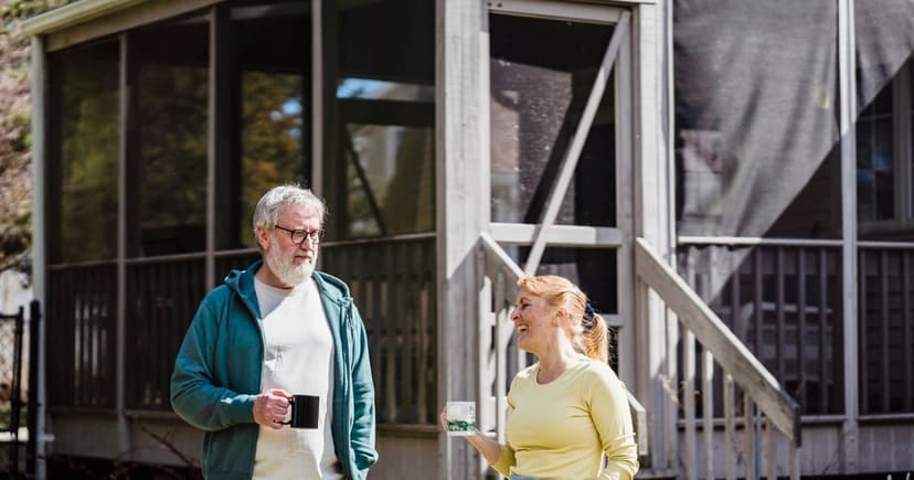A man in a teal zip hoodie and a woman in a yellow top stand talking with coffee mugs in hand outside a weathered screened porch.