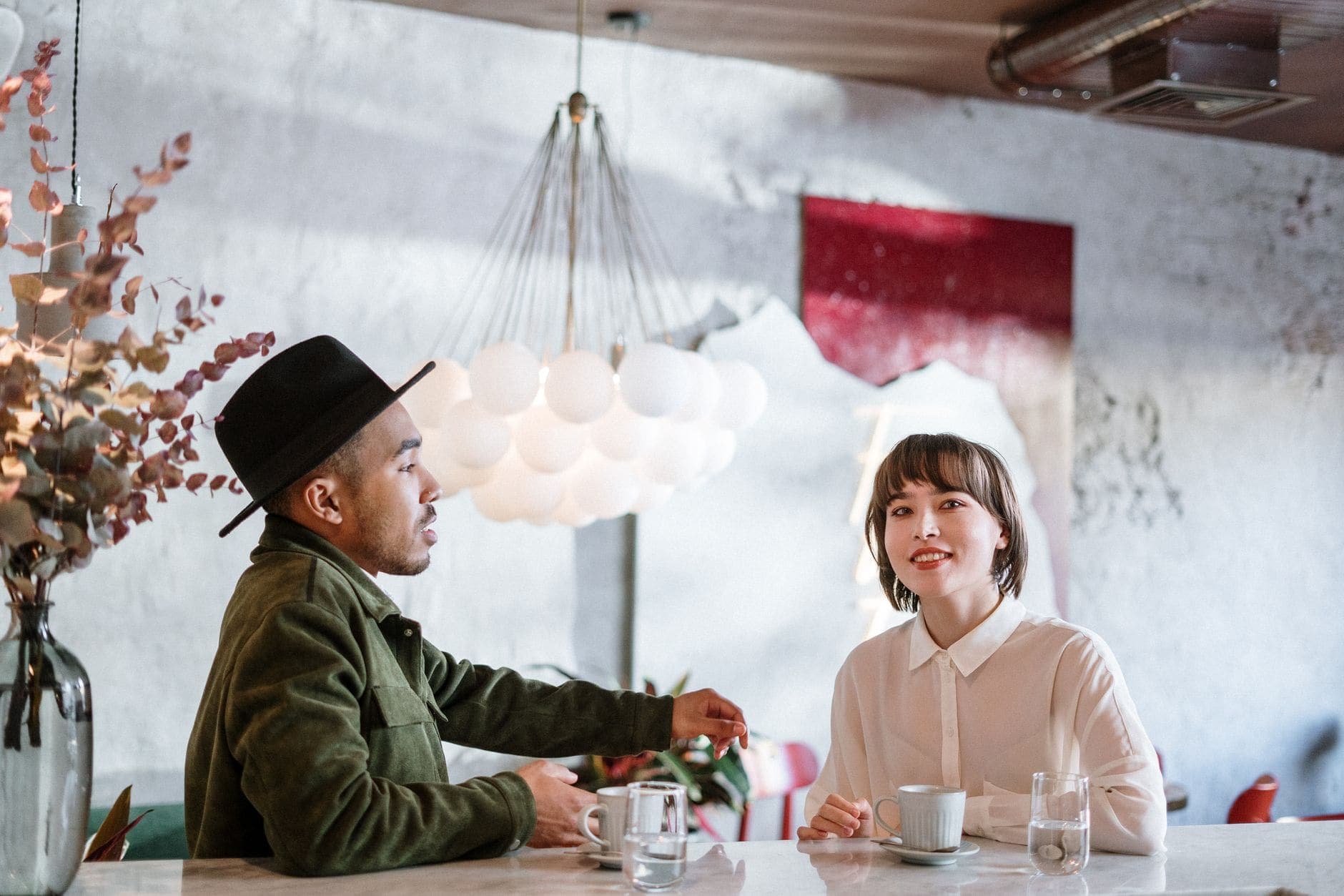 Two adults sit at a table with cups and water glasses under hanging lights.