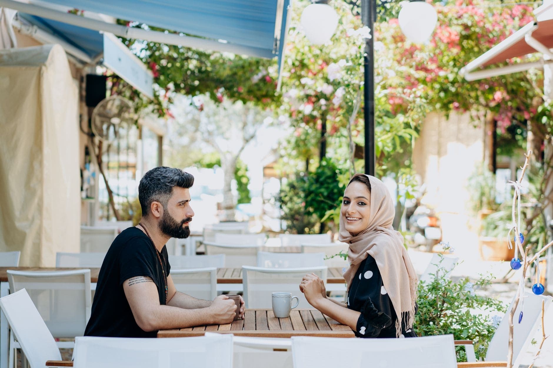 Δύο ενήλικες κάθονται με κούπες σε υπαίθριο καφέ. Η γυναίκα χαμογελά προς την κάμερα.