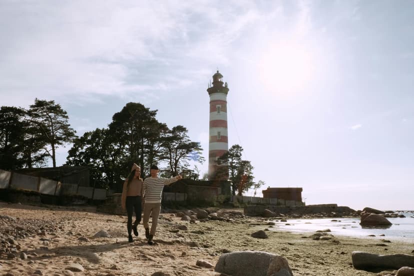 A couple walking hand-in-hand along a sunny coastal beach near a lighthouse