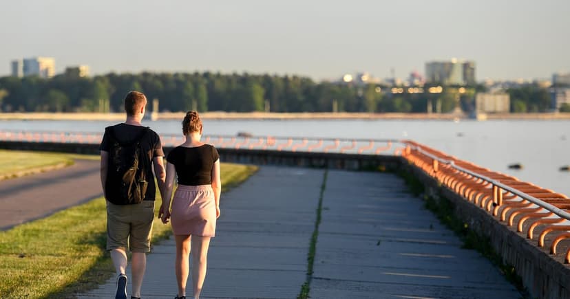 Una pareja pasea de la mano por un paseo marítimo a última hora de la tarde, con la ciudad al otro lado del agua.