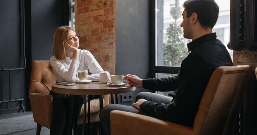 Una pareja conversa en una mesa junto al ventanal de una cafetería de ladrillo visto, con dos cafés sobre la mesa.