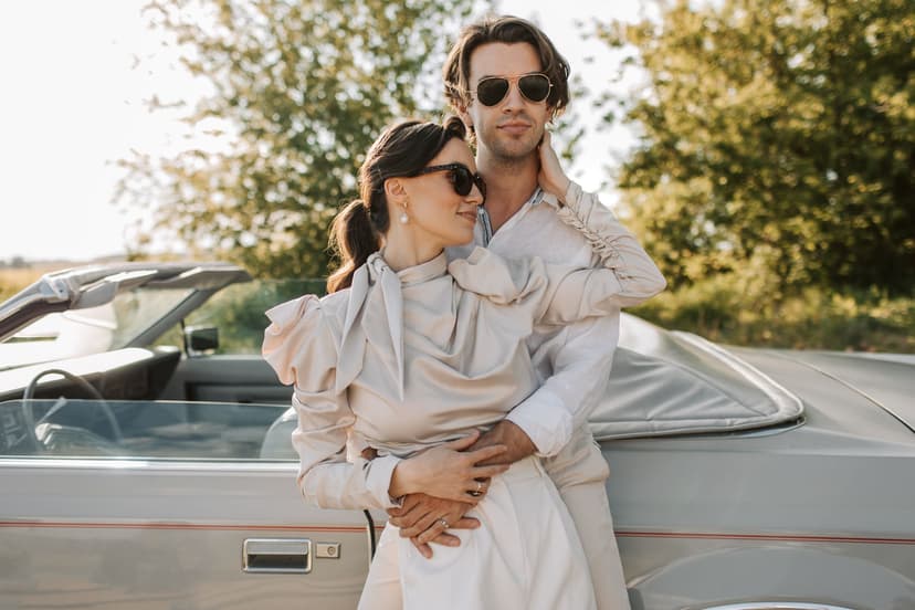 Ein elegant gekleidetes Paar in hellen Sommeroutfits lehnt eng umschlungen an einem silbernen Oldtimer-Cabrio im warmen Abendlicht.