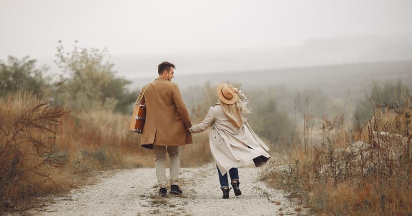 A man in a camel overcoat carrying a guitar case and a woman in a pale trench coat and wide brimmed hat walking away down a gravel road, dry grass on both sides and a flat misty horizon ahead