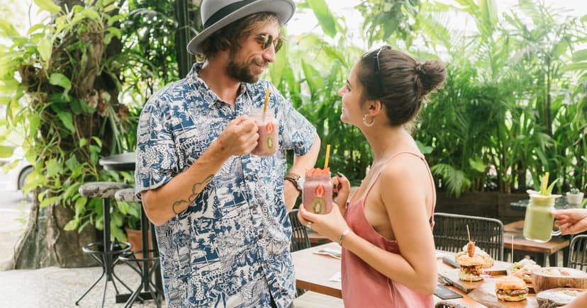 A man in a patterned aloha shirt and a straw hat stands talking with a woman in a coral slip dress, both holding fruit drinks at an open-air table surrounded by palms.