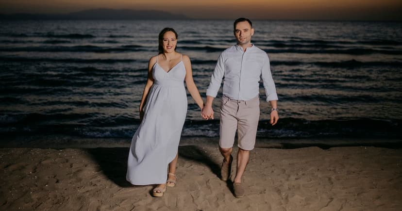 A woman in a long pale dress and a man in a white shirt and shorts walk hand in hand along an empty stretch of sand after sunset, with a dark headland on the horizon