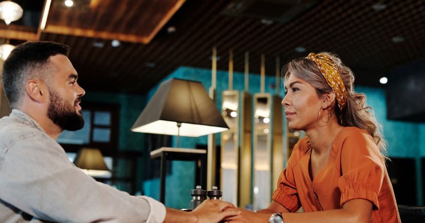 A man in a pale striped shirt and a woman in a rust-orange blouse sit facing each other across a small table in a warmly lit restaurant, their hands resting together.