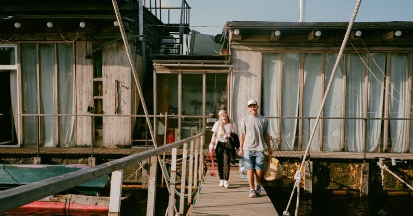 A man in a cap and denim shorts and a woman in a white T-shirt walk a wooden dock past weathered boathouses on a bright summer day.