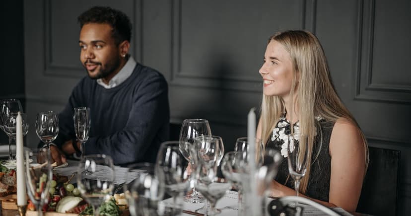 A man in a navy sweater and a blonde woman in a black dress smile at a long dinner table set with wine glasses.