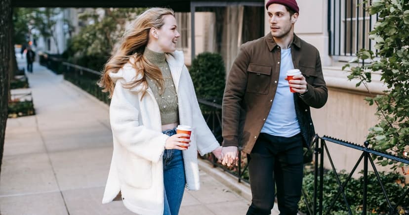 A woman in a cream coat and a man in a brown jacket walk hand in hand along a sidewalk holding takeaway coffee cups.