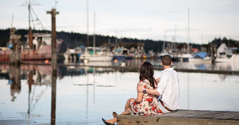 A man with his arm around a woman in a floral dress sits on a wooden dock facing moored fishing boats and calm water.