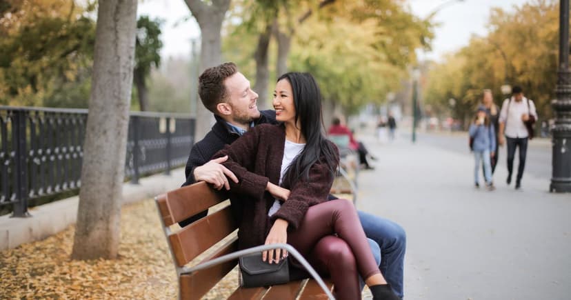 A couple in warm autumn layers sit close on a wooden park bench, laughing together under yellow-leaved trees beside a path.