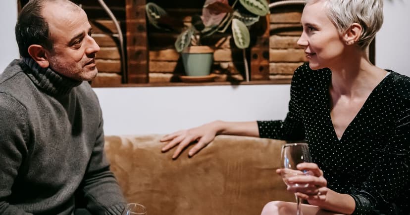 A man in a gray turtleneck and a woman in a black polka-dot dress talk over wine on a sofa.