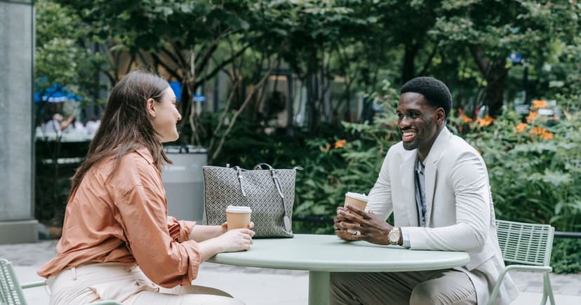 A woman in a rust coloured shirt and a man in a pale linen jacket sit across a small round table outdoors with takeaway coffee cups, other tables behind them.