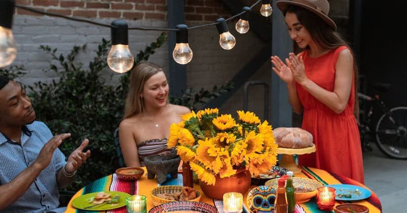A man in a pale blue shirt and a seated blonde woman laugh while a woman in an orange dress stands at an outdoor table under string lights, sunflowers and plates between them.